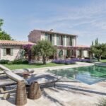 Fachada de piedra con contraventanas verdes, piscina de aguas verdosas, tumbonas y lavanda bajo un cielo azul