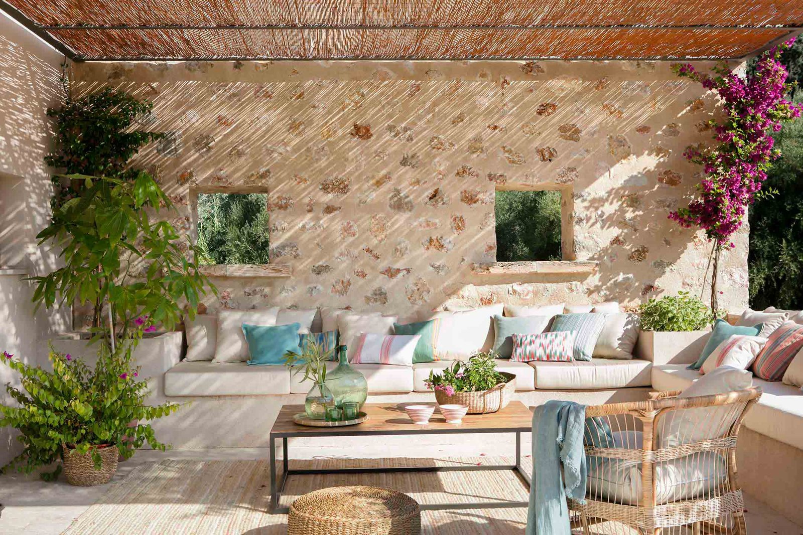 Terraza cubierta con muro de piedra, sofá blanco corrido, cojines de colores y buganvilla