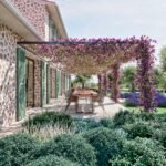 Mesa de comedor exterior bajo pérgola de flores moradas junto a fachada de piedra con persianas verdes