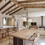 Cocina abierta con isla clara, taburetes de madera, mesa de comedor, chimenea colgante y vigas vistas