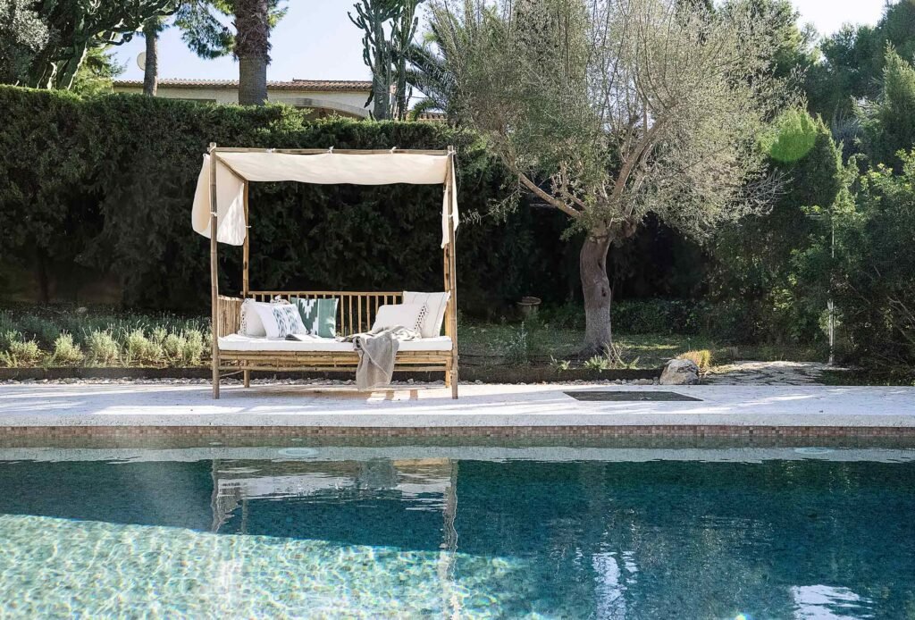Sofá cama de bambú con dosel de tela blanca junto a la piscina y un olivo