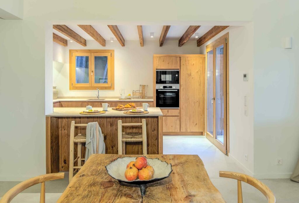 Cocina abierta con barra, armarios de madera natural, vigas vistas y mesa rústica en primer plano