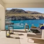 Terraza cubierta con sofá lounge, cojines de colores, linternas y barandilla de cristal frente a la cala