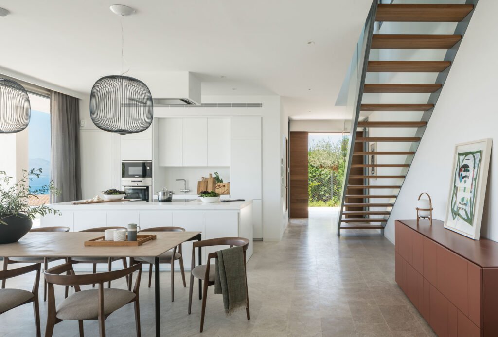 Cocina blanca con isla abierta al comedor, escalera de madera y suelo de piedra clara