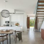 Cocina blanca con isla abierta al comedor, escalera de madera y suelo de piedra clara