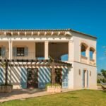 Fachada de dos plantas con terraza cubierta, persianas azules, carpintería de madera y pérgola sobre patio empedrado