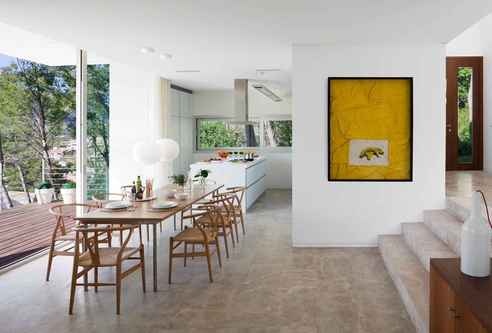 Comedor con mesa y sillas de madera, cocina blanca al fondo, cuadro amarillo y escalones de piedra