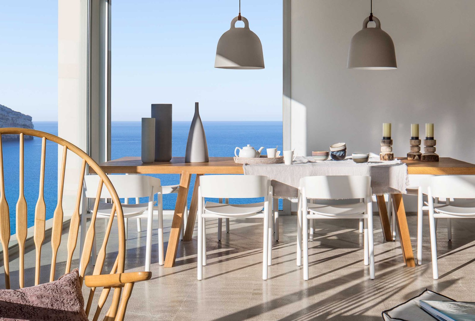 Comedor con mesa de madera, sillas blancas, lámparas colgantes grises y el mar azul al fondo