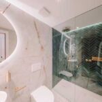 Baño con piedra clara veteada, espejo ovalado iluminado, grifería dorada y ducha de azulejo verde en espiga