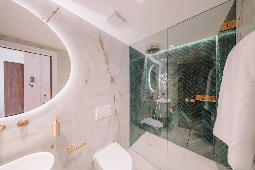 Baño con piedra clara veteada, espejo ovalado iluminado, grifería dorada y ducha de azulejo verde en espiga