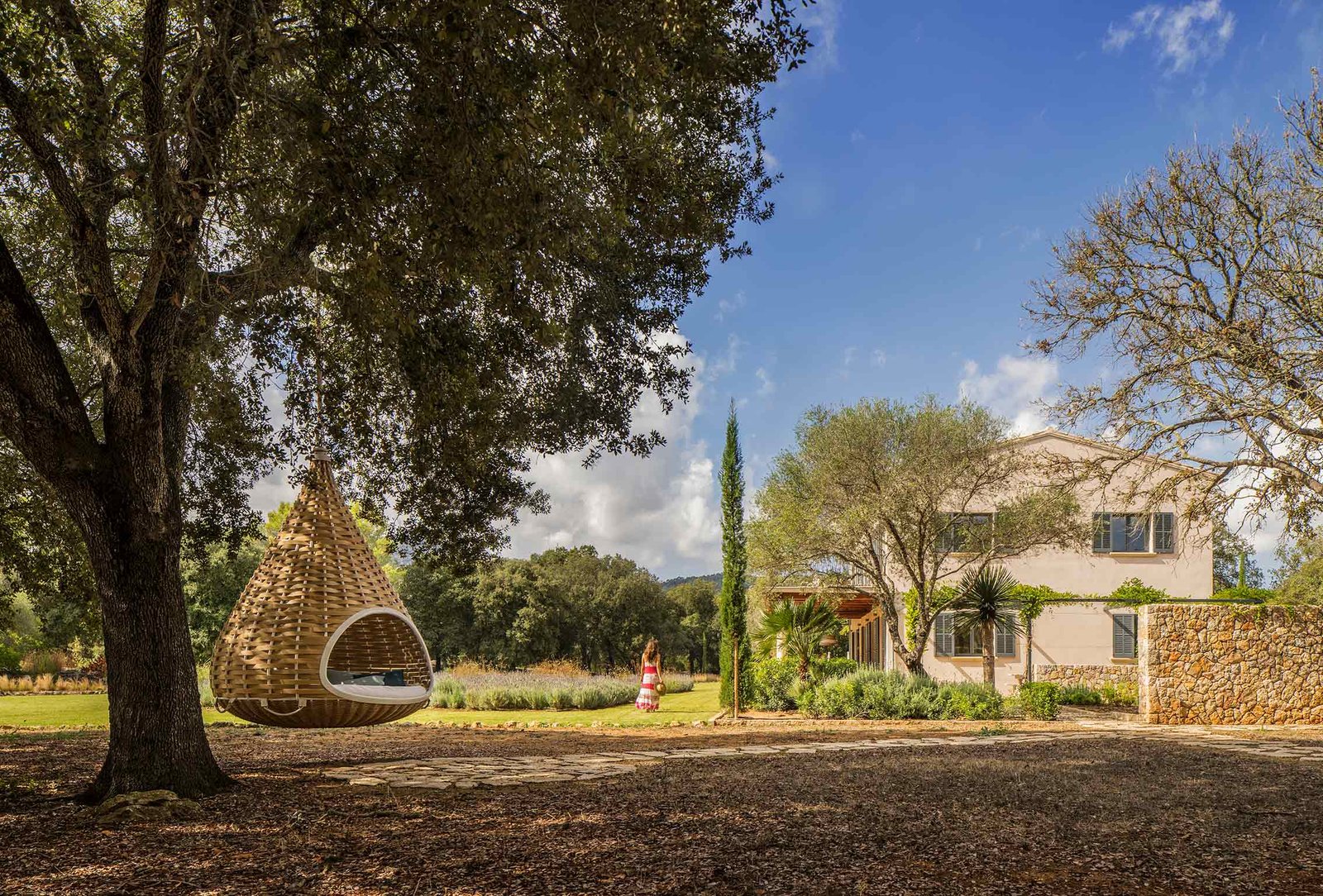 Jardín con cama colgante bajo una encina y la casa al fondo tras un muro de piedra