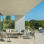 Terraza de piedra clara con sofá, butacas y tumbonas junto a la piscina y monte al fondo