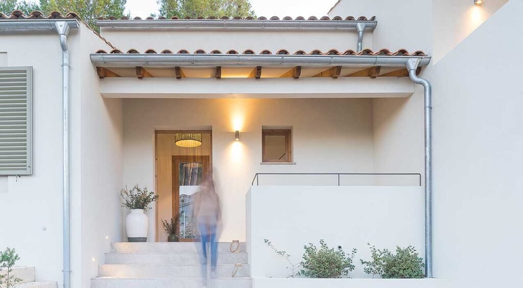 Porche de entrada con puerta de madera, escalera de piedra, tejas, luz cálida y jarrón con plantas