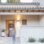 Porche de entrada con puerta de madera, escalera de piedra, tejas, luz cálida y jarrón con plantas