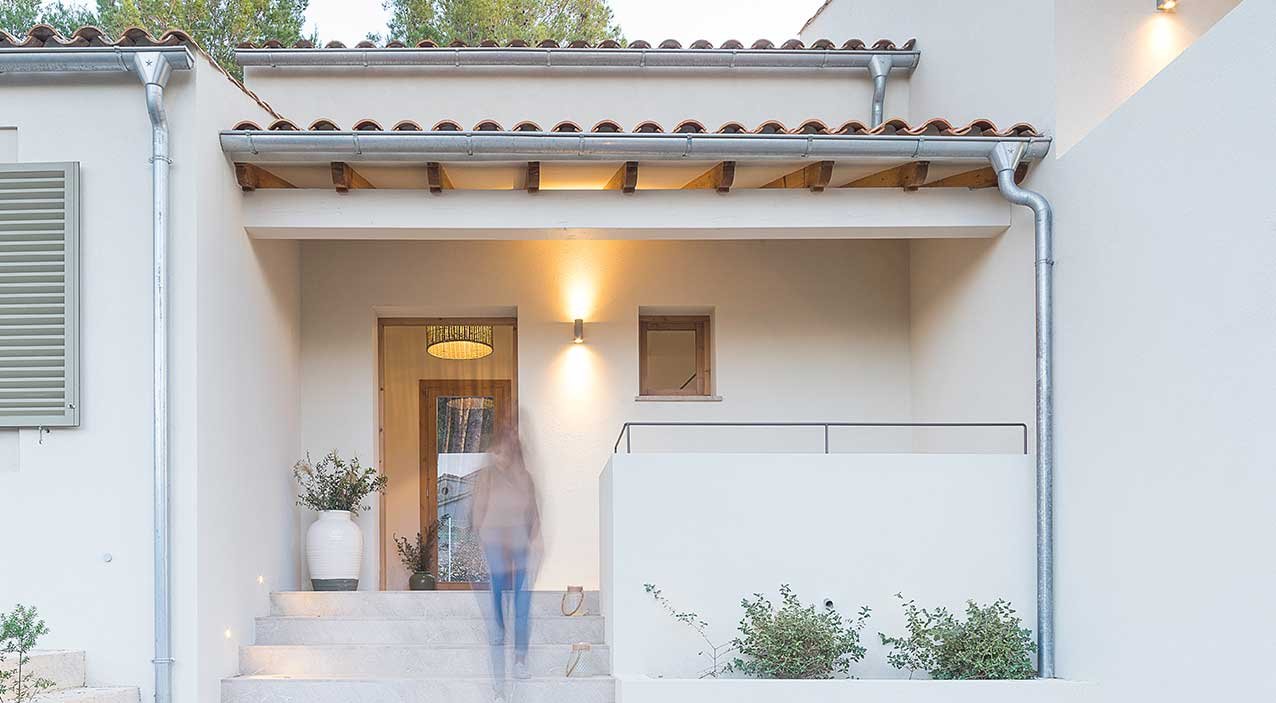 Porche de entrada con puerta de madera, escalera de piedra, tejas, luz cálida y jarrón con plantas