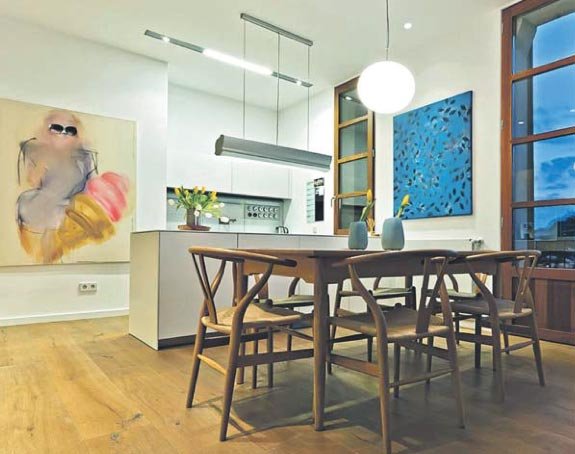 Comedor con mesa y sillas de madera junto a cocina blanca, parqué claro, cuadros abstractos y ventanales