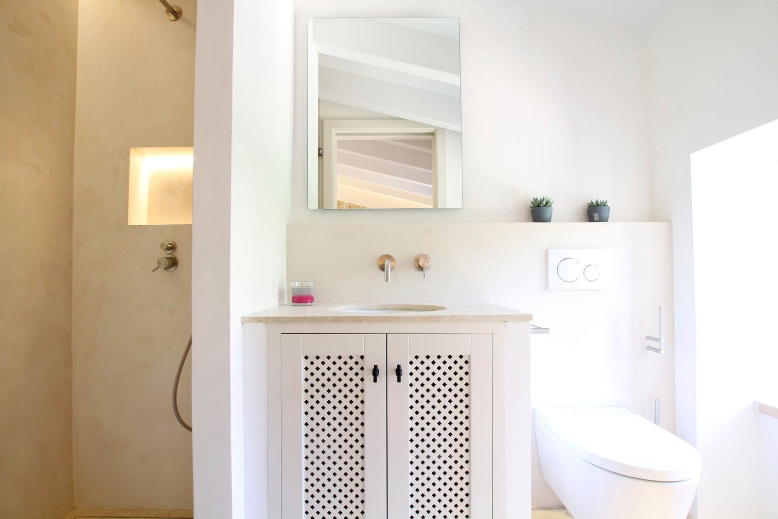 Baño con lavabo sobre mueble blanco de celosía, grifería color cobre, ducha de microcemento y espejo rectangular