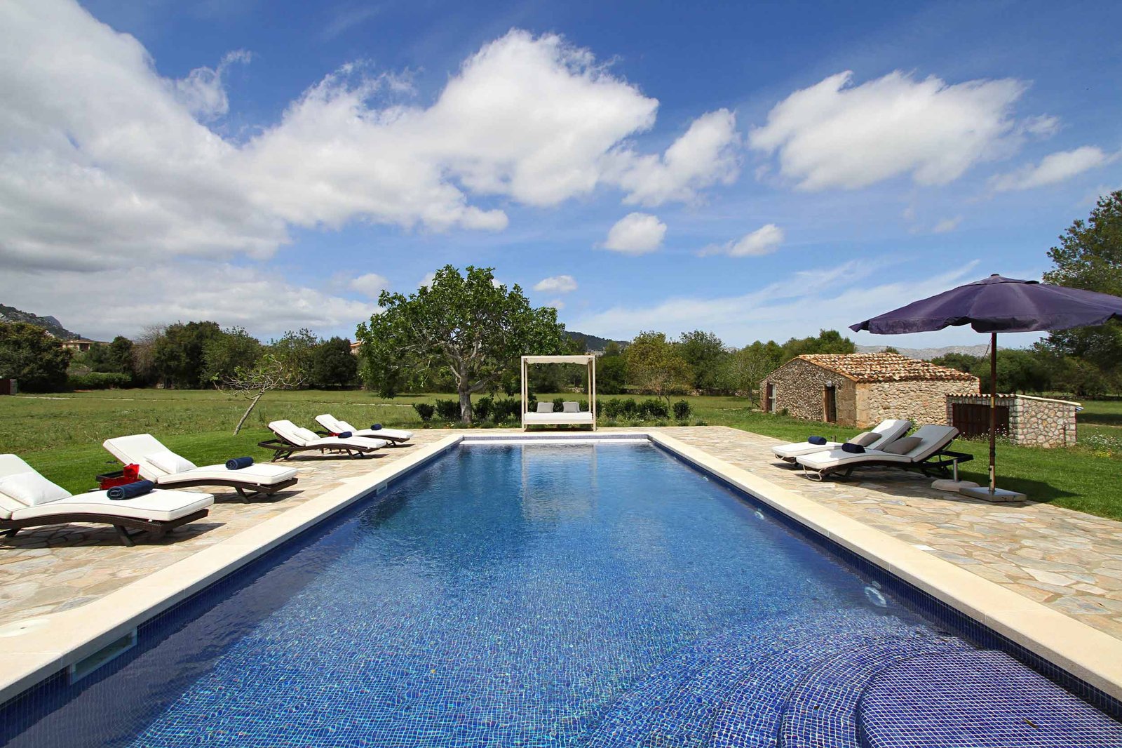 Piscina alargada de mosaico azul con tumbonas, cama con dosel y caseta de piedra entre prados verdes