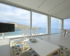 Salón de Cala Carbó con grandes ventanales al mar, sillas blancas, mesa baja y alfombra estampada