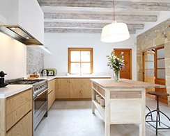 Cocina de Ca'n Barret con muebles de madera clara, isla central, vigas vistas y pared de piedra