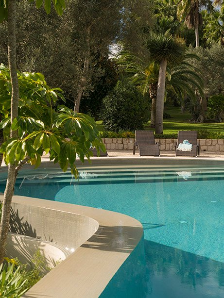 Piscina de agua turquesa con escalones de acceso, tumbonas al fondo, muro de piedra y jardín arbolado