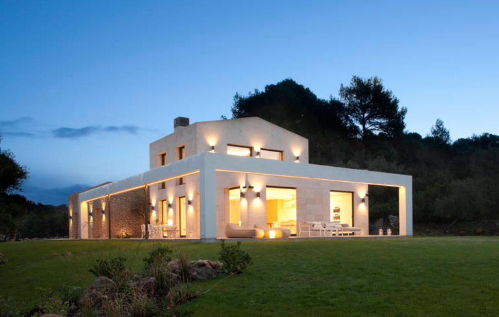 Fachada de la vivienda AC3 al anochecer, con piedra clara, pórtico blanco, luz cálida y césped