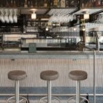 Tres taburetes de acero y asiento marrón frente a la barra, con botellas y copas colgadas