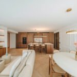 Salón abierto con sofá crema, mesa ovalada con sillas de rejilla y cocina de nogal al fondo