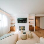 Salón luminoso con visillos blancos, televisión sobre chimenea, estantes de nogal y sofá crema