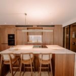 Isla de cocina con encimera de piedra clara, tres taburetes de rejilla, muebles de nogal y vitrina