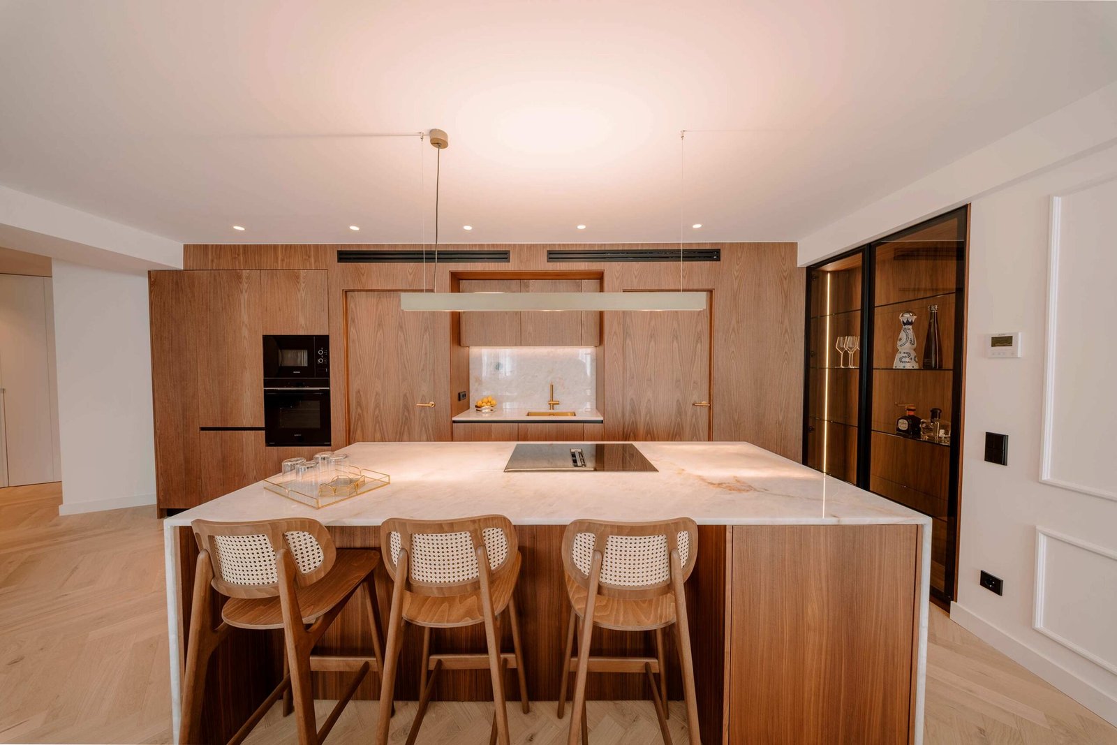 Isla de cocina con encimera de piedra clara, tres taburetes de rejilla, muebles de nogal y vitrina