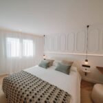 Dormitorio con pared de molduras ovaladas, colcha de cuadros, lámparas colgantes y visillos blancos