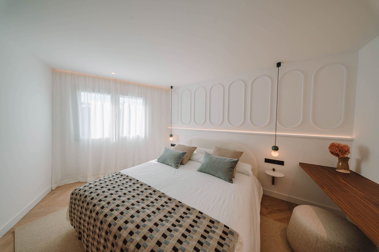 Dormitorio con pared de molduras ovaladas, colcha de cuadros, lámparas colgantes y visillos blancos