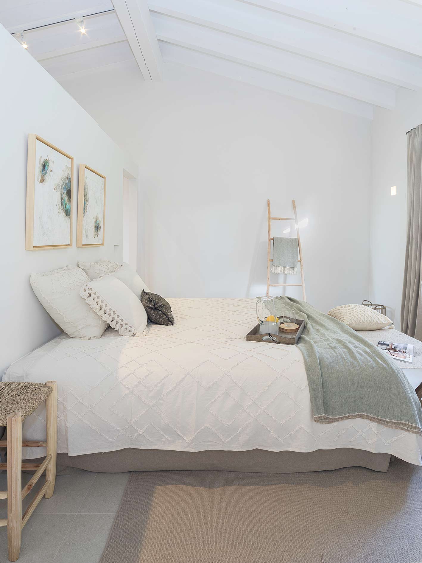 Dormitorio blanco de techo inclinado con vigas, colcha con relieve, manta verde y escalera decorativa