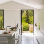Isla de cocina con taburetes y puerta abierta al jardín, con muro de piedra y vegetación