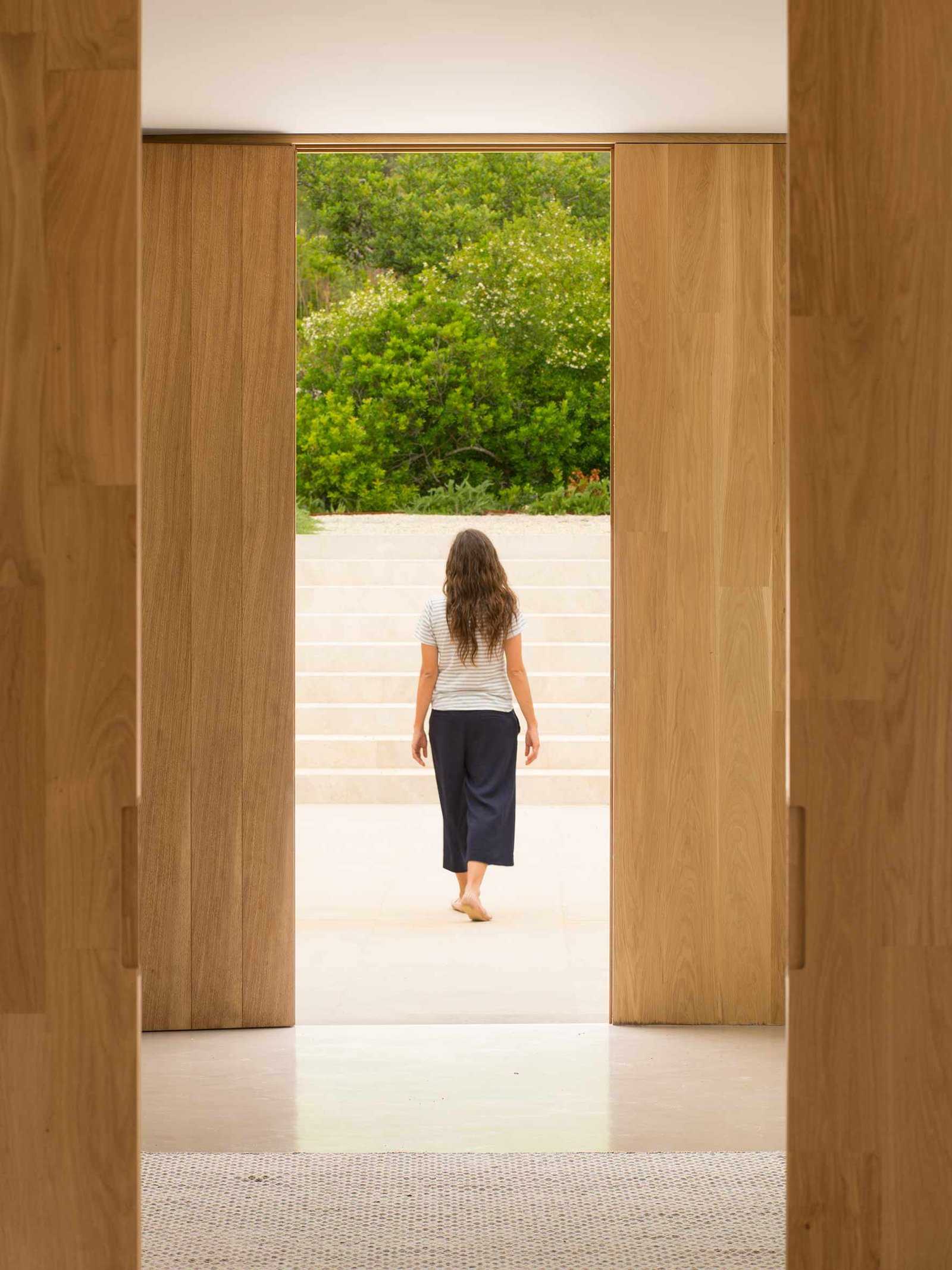 Puertas de roble abiertas hacia una escalinata de piedra clara, con una persona de espaldas y jardín