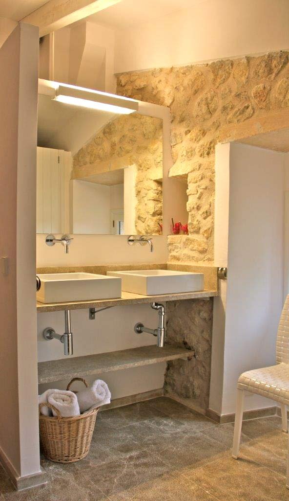 Baño con dos lavabos blancos sobre encimera de piedra, pared de piedra vista y cesta con toallas