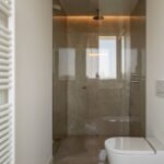 Ducha acristalada con revestimiento veteado color topo, luz indirecta, inodoro suspendido y radiador toallero