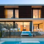 Fachada de la casa LF115 al anochecer con lamas de madera, piscina y terraza iluminada