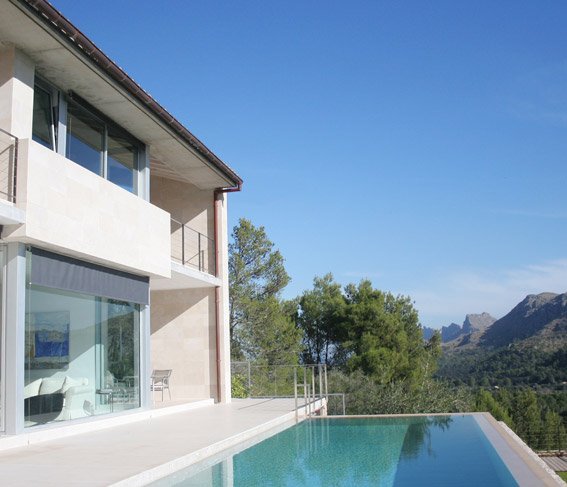 Vivienda LF96 con fachada de piedra clara, grandes cristaleras, terraza y piscina infinita frente a la montaña