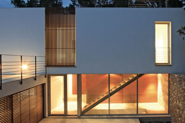 Fachada blanca de Blanca pureza al anochecer con lamas de madera, cristalera iluminada y muro de piedra