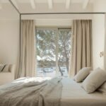 Dormitorio con cama de dosel, cortinas de lino beige y vistas a una bahía con barcas