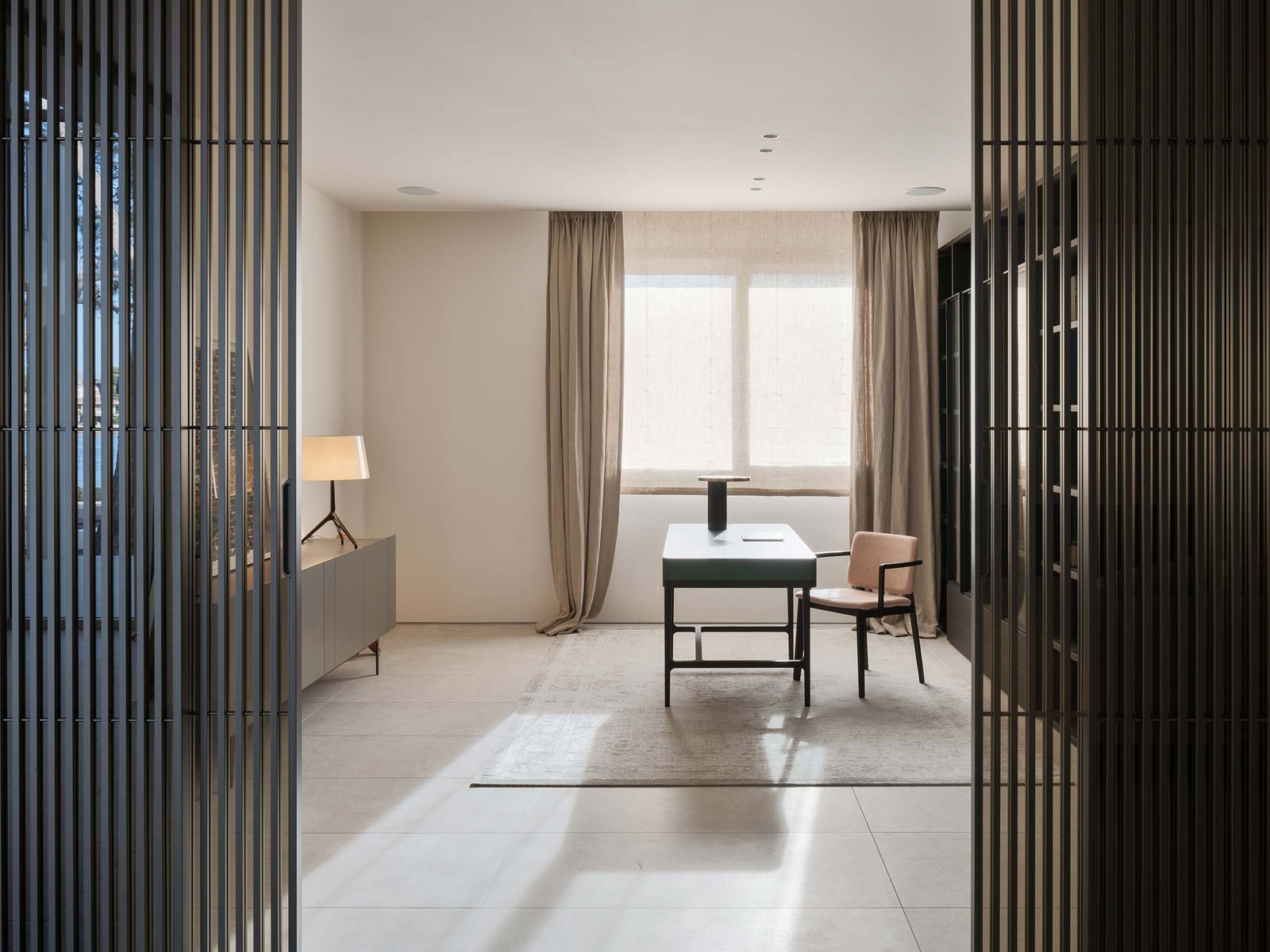 Despacho con escritorio verde, silla rosa, aparador y cortinas de lino tras lamas de madera oscura