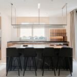 Cocina con isla de mármol oscuro, cuatro taburetes negros, armarios de madera y estante colgante