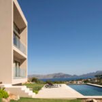 Fachada beige con balcones de cristal junto a piscina y jardín, con mar y montañas al fondo
