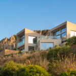 Villa escalonada en la ladera rocosa vista desde abajo, con volúmenes beige y barandillas de cristal