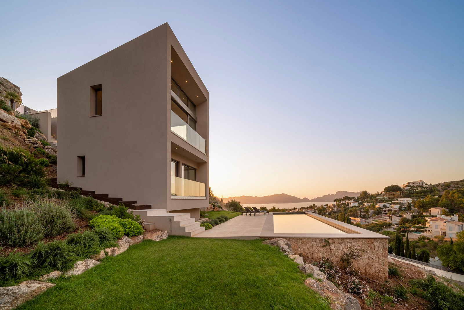 Volumen de fachada beige al atardecer con jardín de césped, piscina y vistas a la bahía