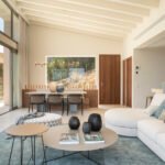 Salón comedor de planta abierta con vigas blancas, sofá curvo, mesa de comedor y cocina al fondo