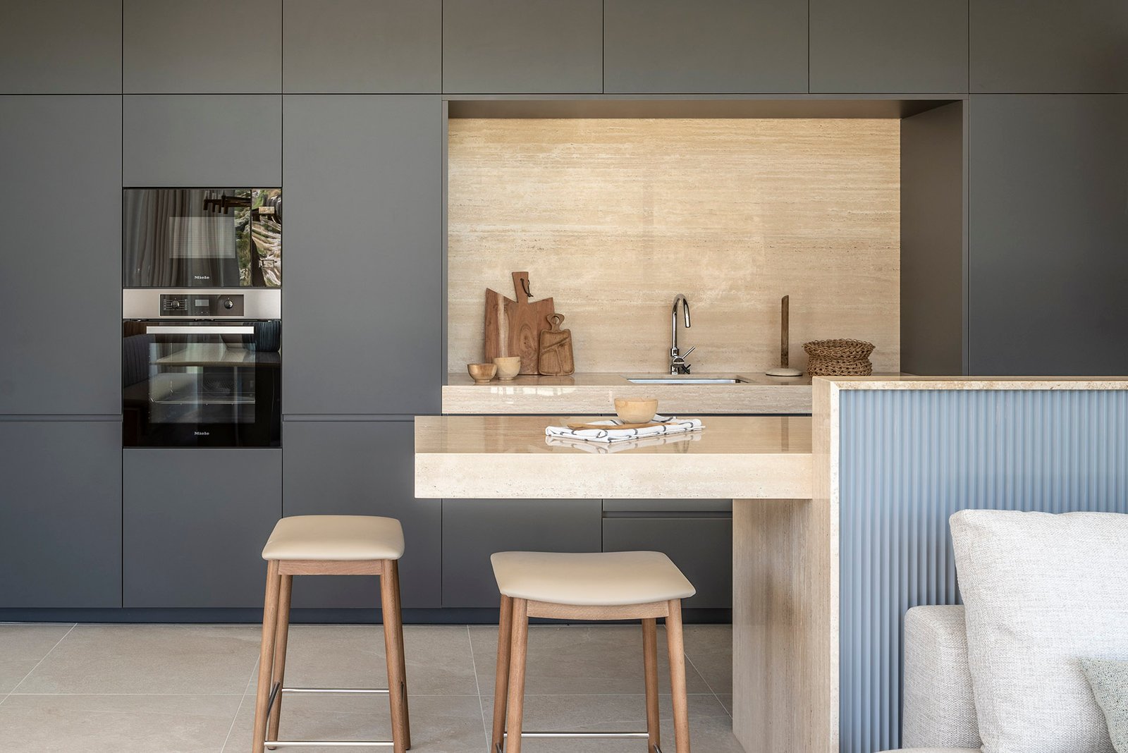 Cocina con armarios gris oscuro, isla y frontal de piedra beige veteada y taburetes de madera