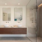 Ducha con mampara de cristal y revestimiento gris, junto a espejos rectangulares y grifería de pared